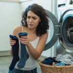 Frustrated woman holding different socks in front of a washing machine with a laundry basket nearby.