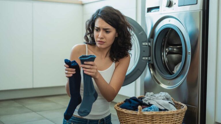 Frustrated woman holding different socks in front of a washing machine with a laundry basket nearby.