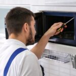 A person in blue overalls uses a tool to work on a microwave oven in a kitchen setting.
