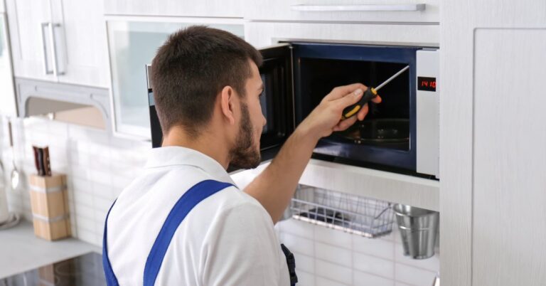 A person in blue overalls uses a tool to work on a microwave oven in a kitchen setting.