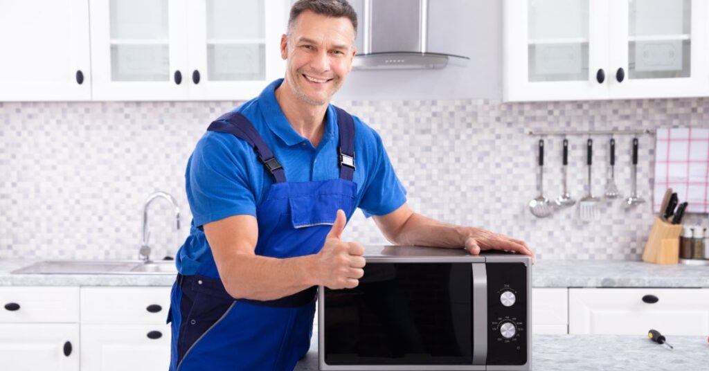 A technician in a blue uniform stands in a modern kitchen, giving a thumbs-up while standing next to a microwave on the counter.