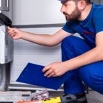 A technician in blue overalls repairs a refrigerator leaking water, surrounded by tools and a red toolbox.