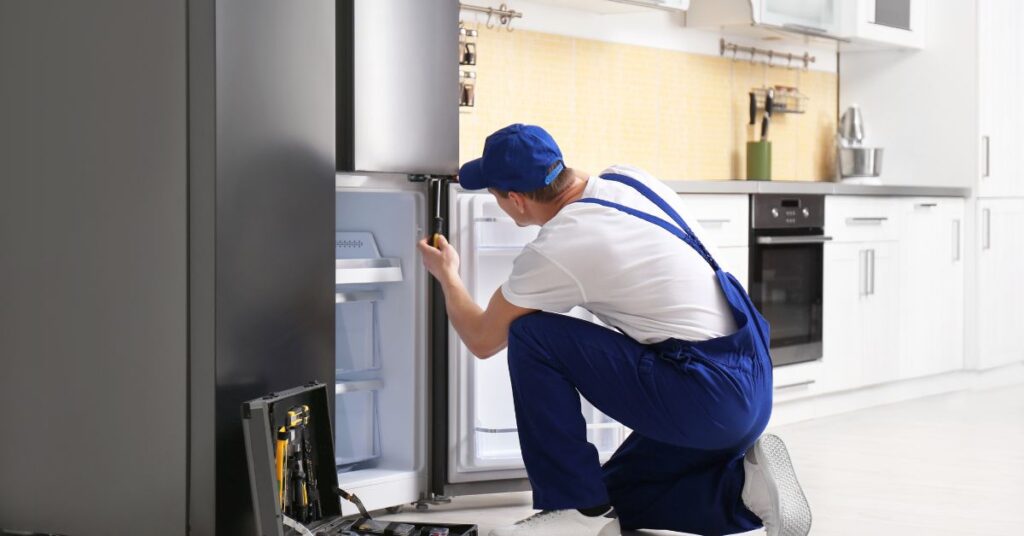 A technician in a blue jumpsuit and cap is kneeling in front of an open refrigerator in a modern kitchen, appearing to inspect or repair it.